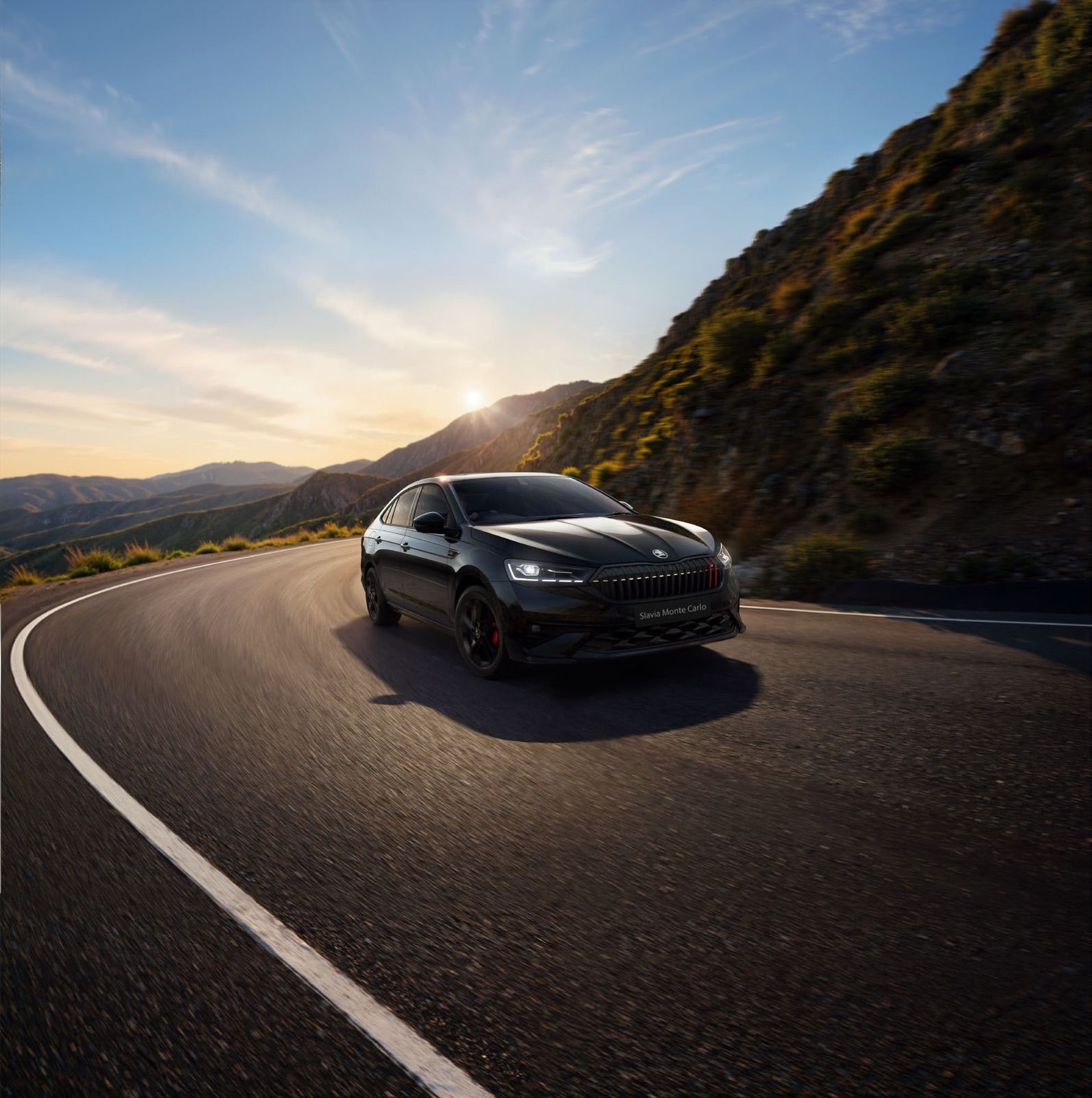 2026 Škoda Slavia Monte Carlo facelift driving on a mountain road at sunset with LED headlamps illuminated
