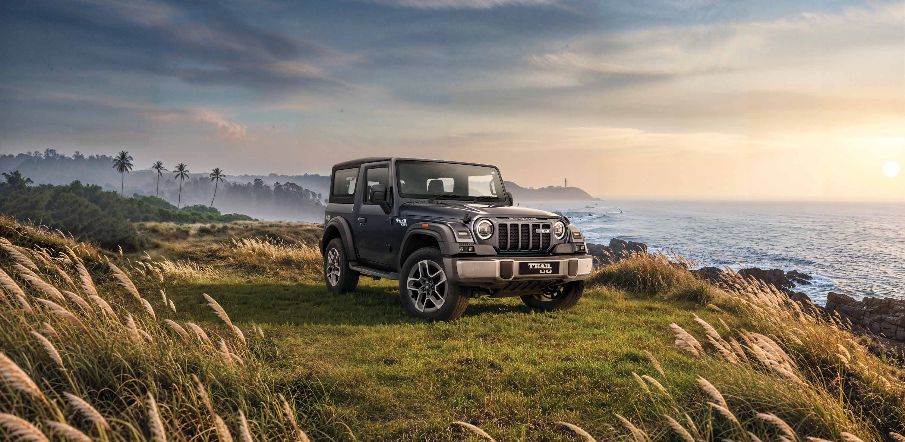 Mahindra Thar OG, Dark grey Thar OG parked in tall coastal grass on a headland at sunrise or sunset, sea and rocks to the right, palms and